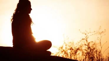A silhouette of a person sitting in meditation pose on a hilltop at sunset, golden light glowing behind them through tall grasses.