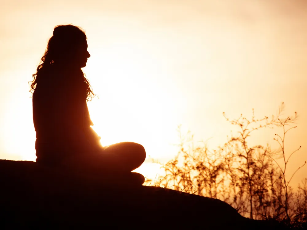 A silhouette of a person sitting in meditation pose on a hilltop at sunset, golden light glowing behind them through tall grasses.