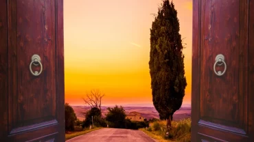 Two tall wooden doors swung open to reveal a quiet country road leading toward a hillside farmhouse under a glowing orange sunset sky.