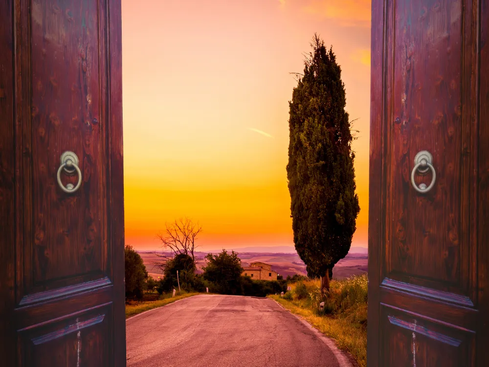Two tall wooden doors swung open to reveal a quiet country road leading toward a hillside farmhouse under a glowing orange sunset sky.