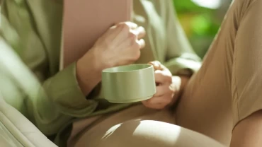 A person sitting in a soft chair holding a green mug and a book, in warm natural light — a quiet moment of contemplation.