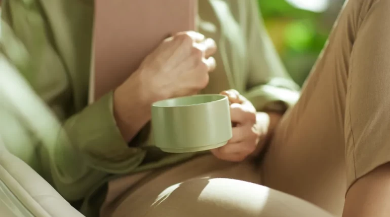 A person sitting in a soft chair holding a green mug and a book, in warm natural light — a quiet moment of contemplation.