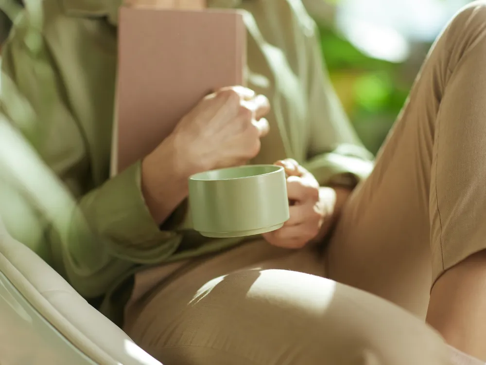 A person sitting in a soft chair holding a green mug and a book, in warm natural light — a quiet moment of contemplation.