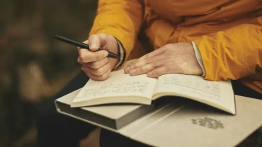 A person in a mustard-yellow jacket sitting outdoors, writing with a black pen in an open notebook resting on their lap.