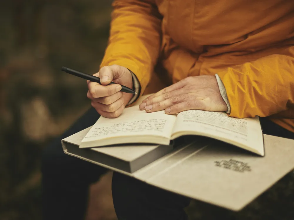 A person in a mustard-yellow jacket sitting outdoors, writing with a black pen in an open notebook resting on their lap.